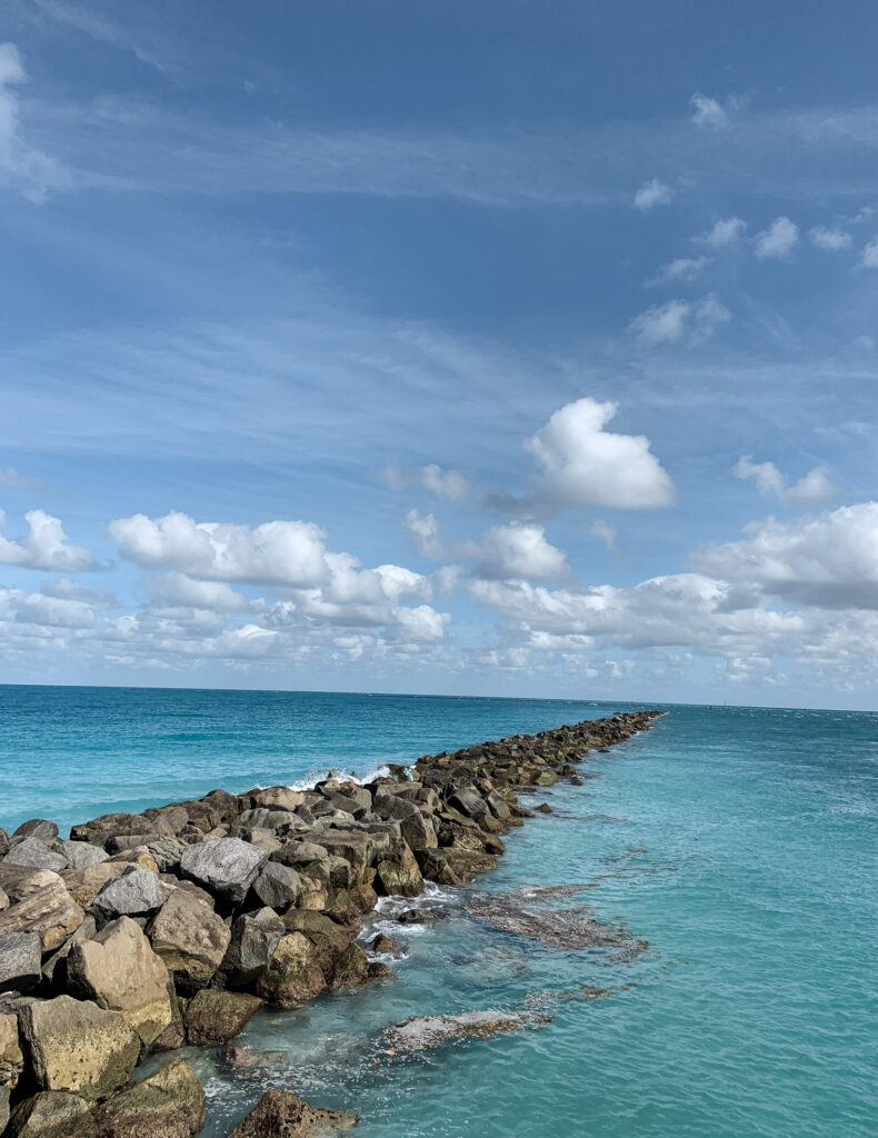 A rock jetty extends into the distance in a quiet ocean under a blue sky