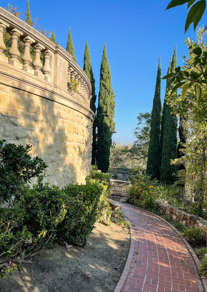 Curved brick garden path beside a stone balustrade and tall cypress trees in warm Mediterranean light.