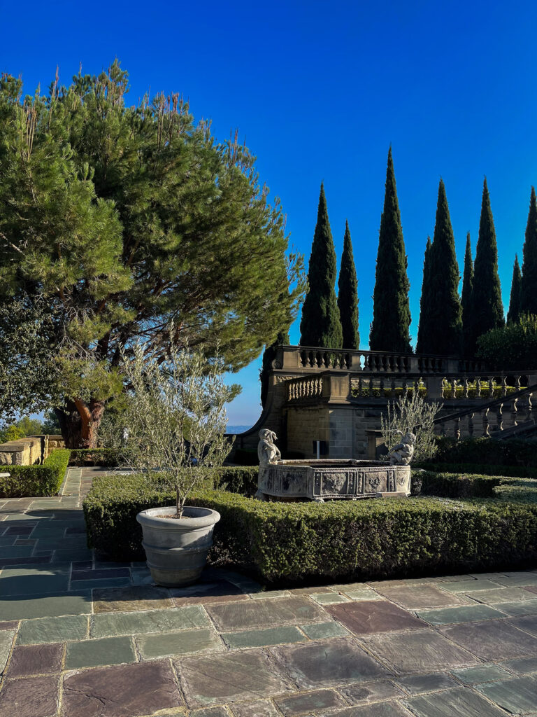 A fountain and hedges sit in a stone courtyard with conifer and oak trees behind a stone balustrade and stairs.