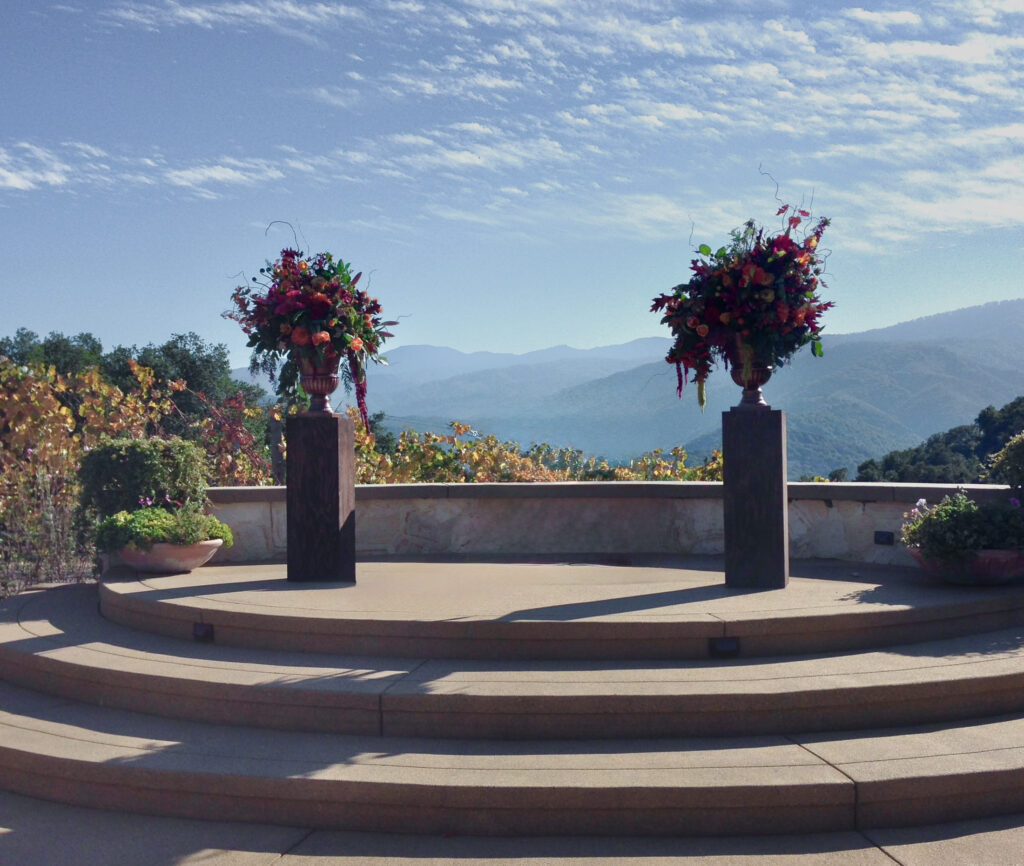Sunlit courtyard steps overlook distant mountains. Flower arrangements on pedastals stand on the ledge. Fall foliage surrounds.
