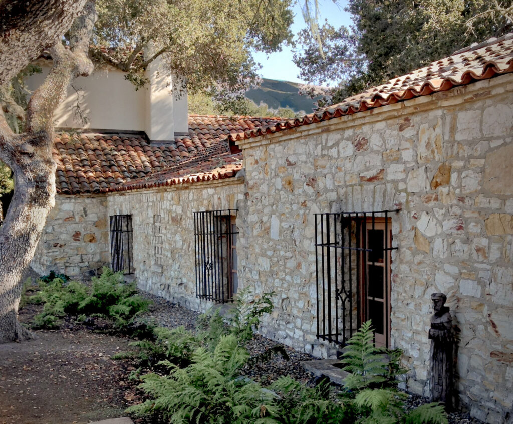 A stone Mediterranean house with an orange tile roof is seen on an angle. A statue of St. Francis is along the wall and green ferns are on the ground along with a large oak tree.