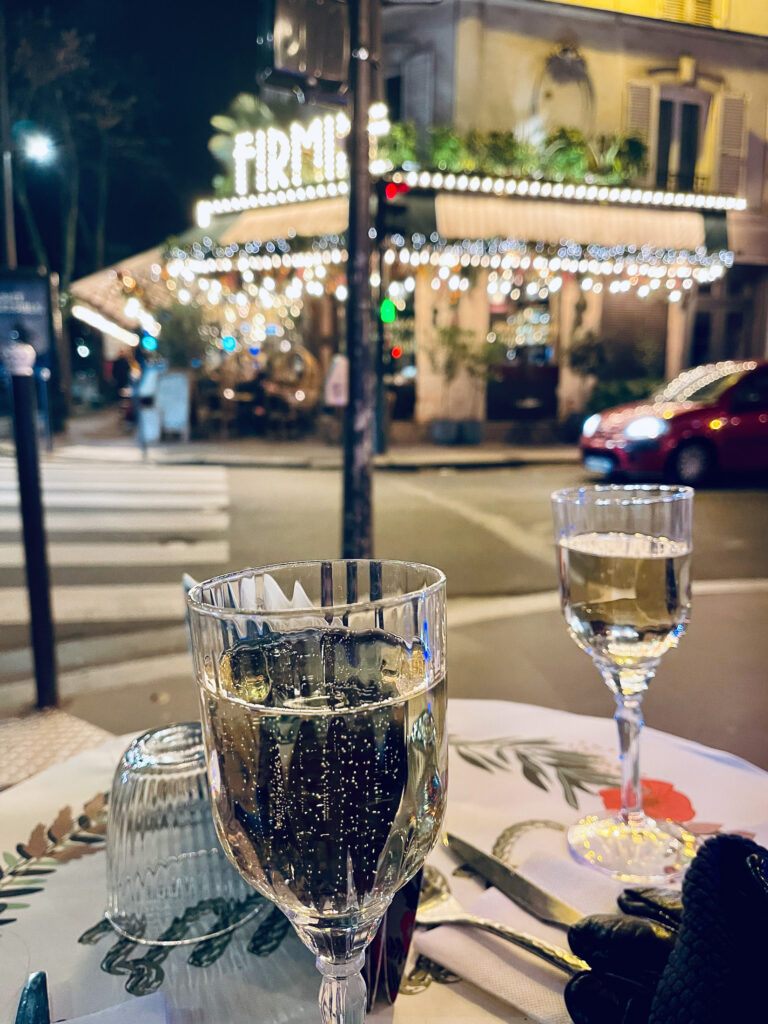 At nighttime, two glasses filled with sparkling champagne sit on a white tablecloth on a corner table on a French Paris street corner. Across the street another cafe has sparkling hanging lights.