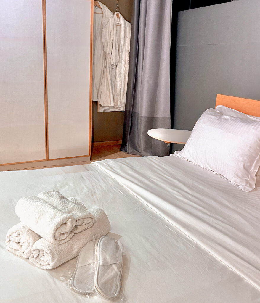 A bed with crisp white linens sits foreground, with white robes hanging on hangers near a white armoire in background. On the bed are rolled towels and hotel slippers.