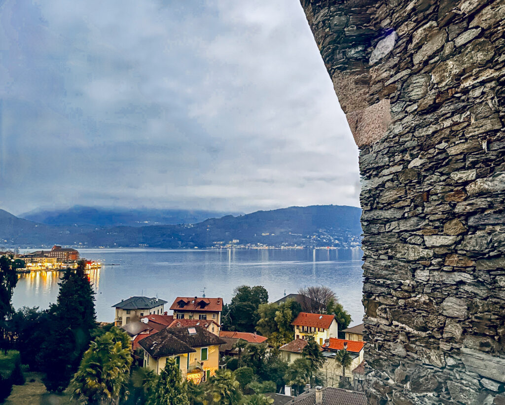View through a castle window overlooking a calm peaceful lake at dusk with foggy cloudy mountains in the distance. Foreground is a village with white buildings and red orange tile roofs, foliage and trees.