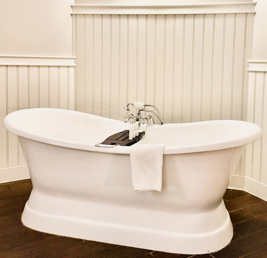 White bathtub with wooden tray and hand towel sit on a brown wooden floor with white beadboard wall behind