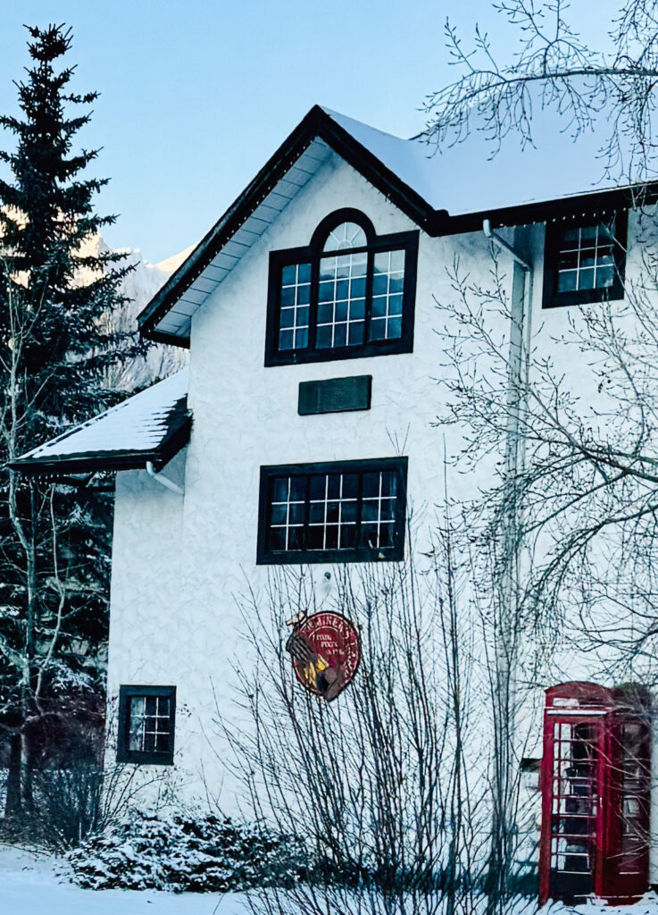 Snow-covered alpine lodge with steep rooflines, tall evergreen tree, and red phone booth in quiet winter setting
