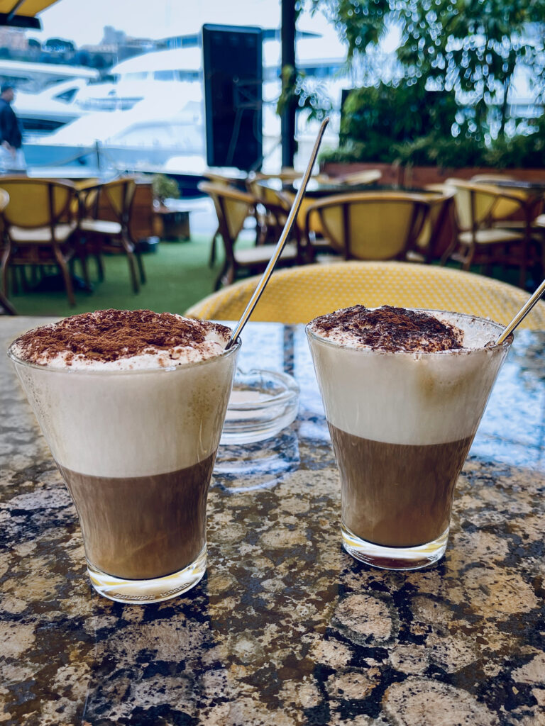 Closeup view of two frothy cappuccinos in glasses, topped with shaved chocolate and cinnamon. The glasses sit on a gleaming granite countertop, and we see yachts in the harbor just outside.