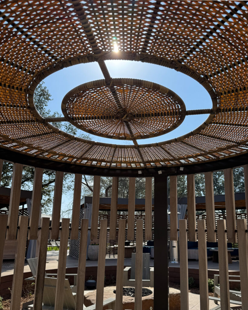 Architectural shot of woven wooden cabana roof with sun shining through and lounge chairs with fire pit beyond