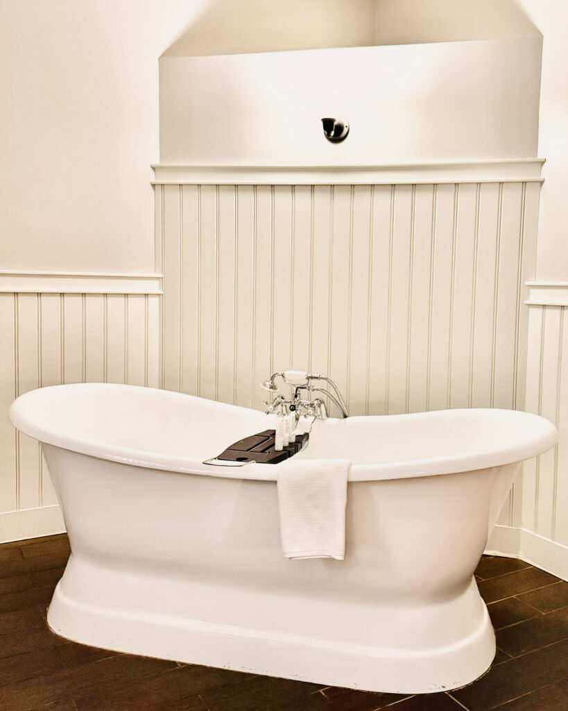 White bathtub with wooden tray and hand towel sit on a brown wooden floor with white beadboard wall behind