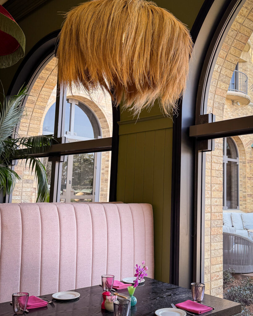 A black table next to an arched window is set for four with a pink velvet settee and feathered chandelier overhead