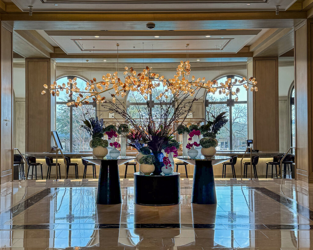 Floral arrangements stand tall on marble pedastals in a grand lobby with chandelier and arched windows behind