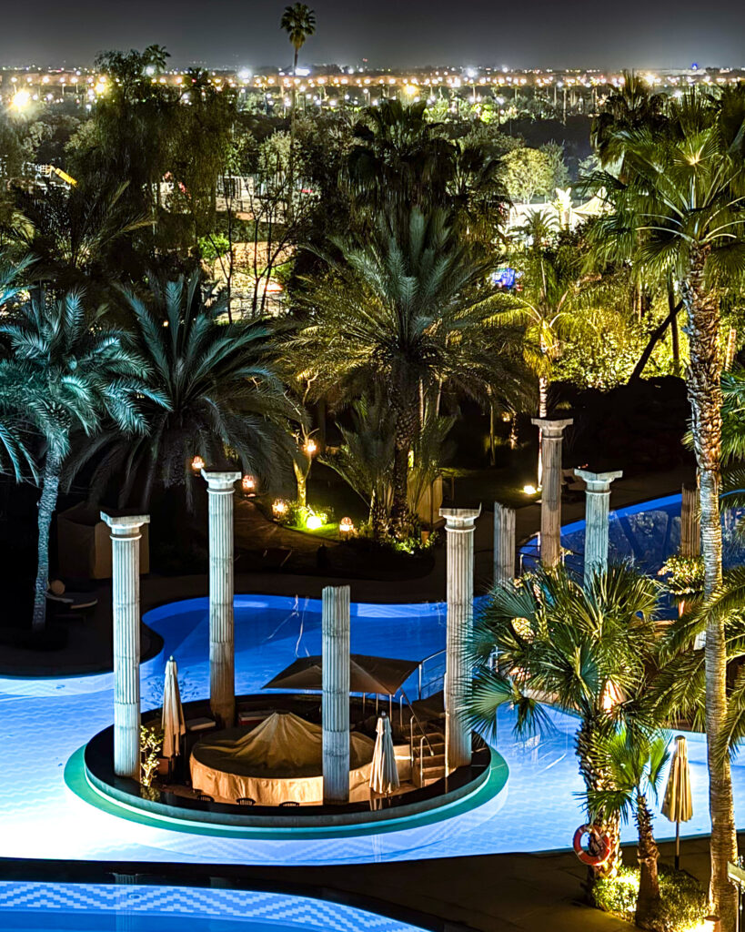 Night view of pool with island in the middle with columns, surrounded by palm trees with city lights in distance